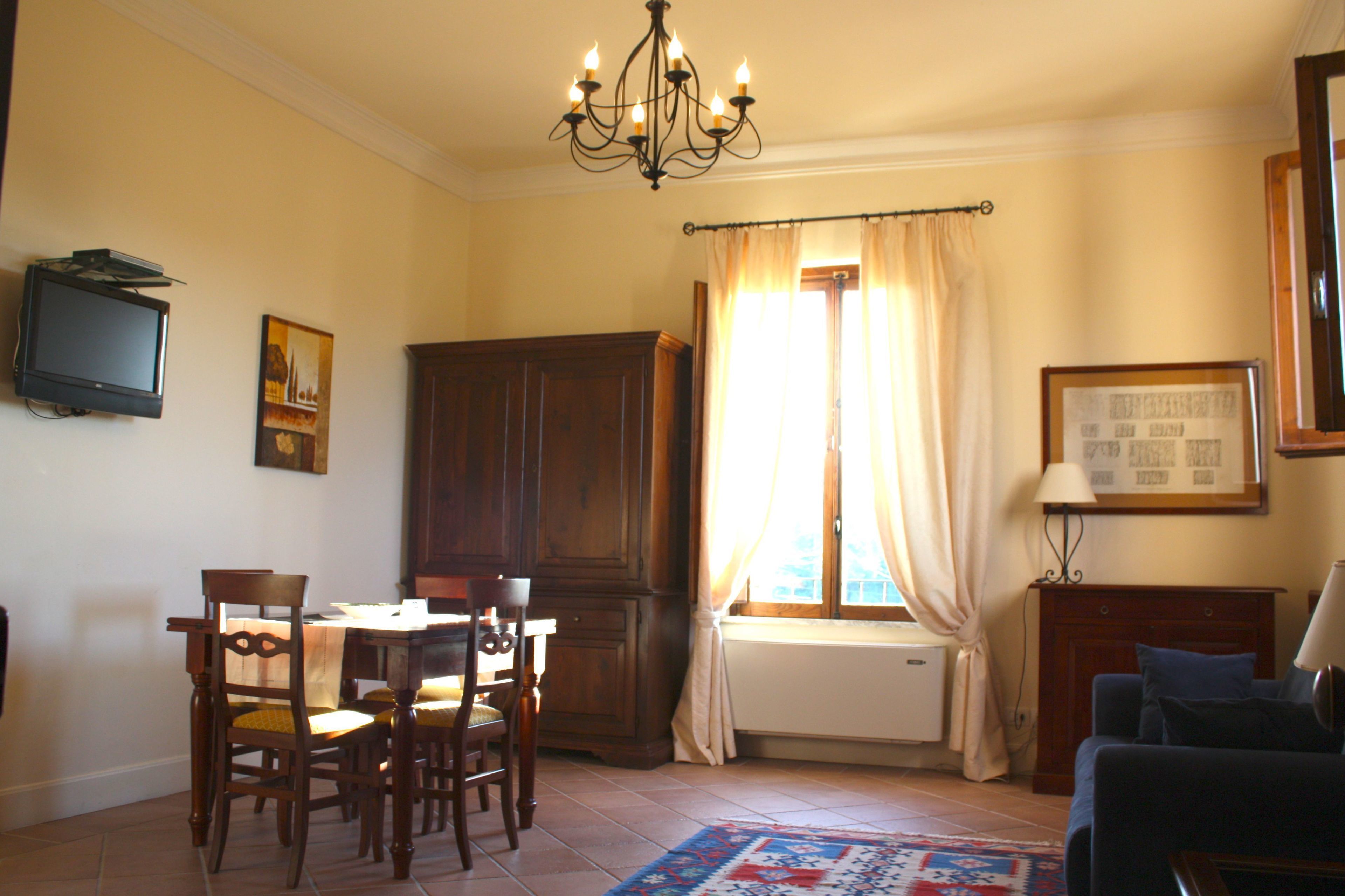 A living room with a dining area, a television mounted on the wall, a chandelier, and a window with curtains.