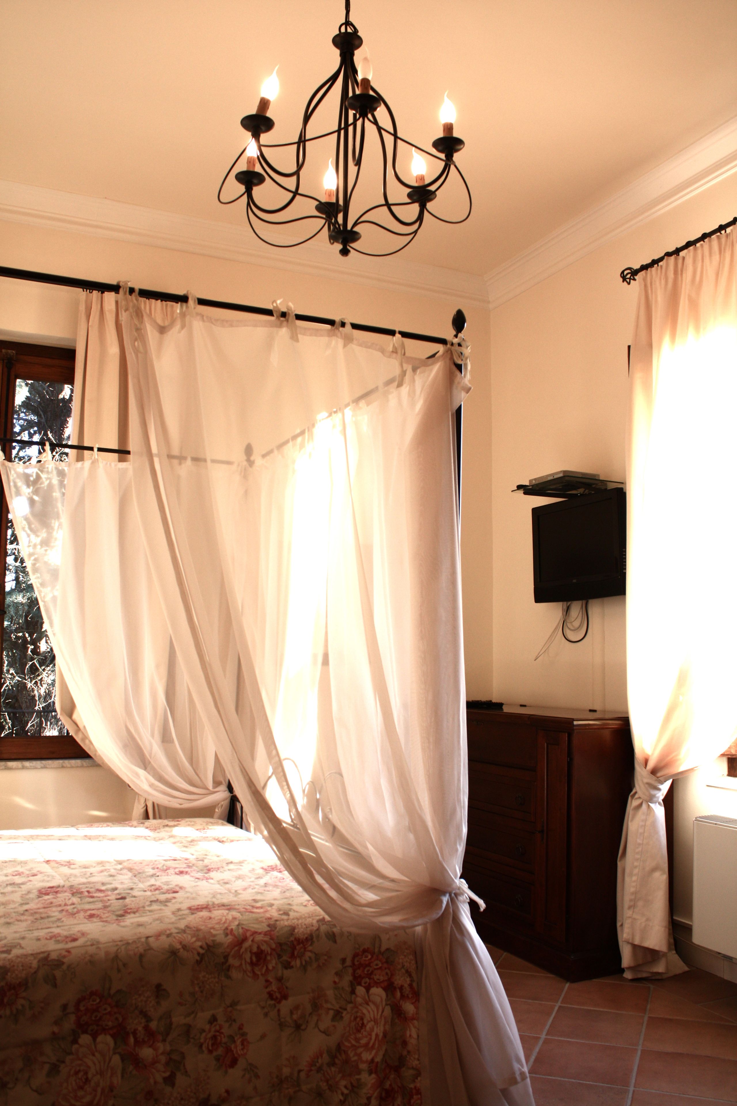 Ein Schlafzimmer mit Himmelbett, einer Kronleuchter, einem an der Wand befestigten Fernseher und einem Fenster mit Vorhängen.