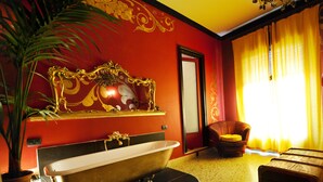 Un baño con una bañera independiente, una silla con cojín, una planta en maceta y un espejo con detalles ornamentales dorados.