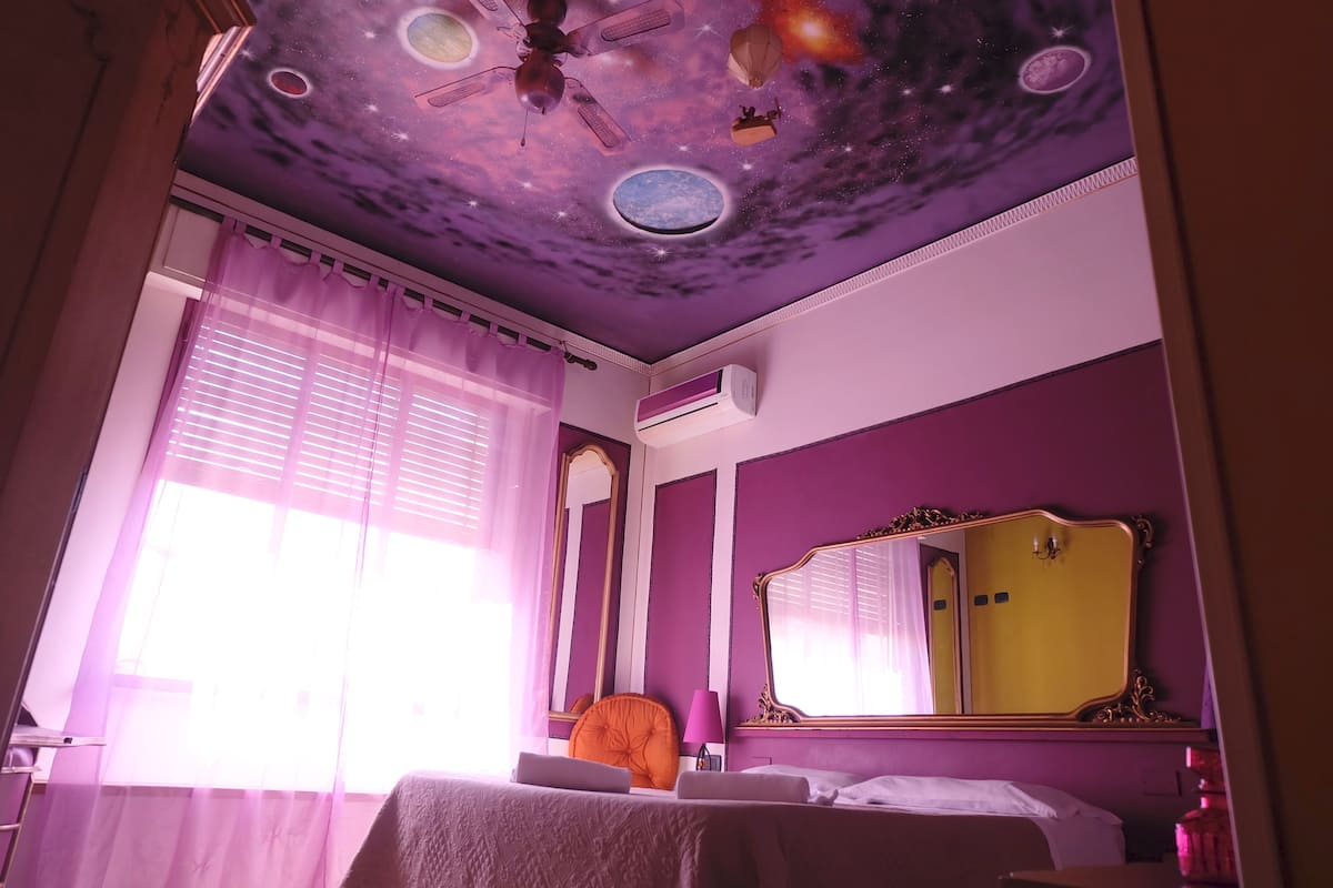 Una habitación con temática morada, una cama, un escritorio, una silla, un techo con un mural al espacio y un espejo grande.