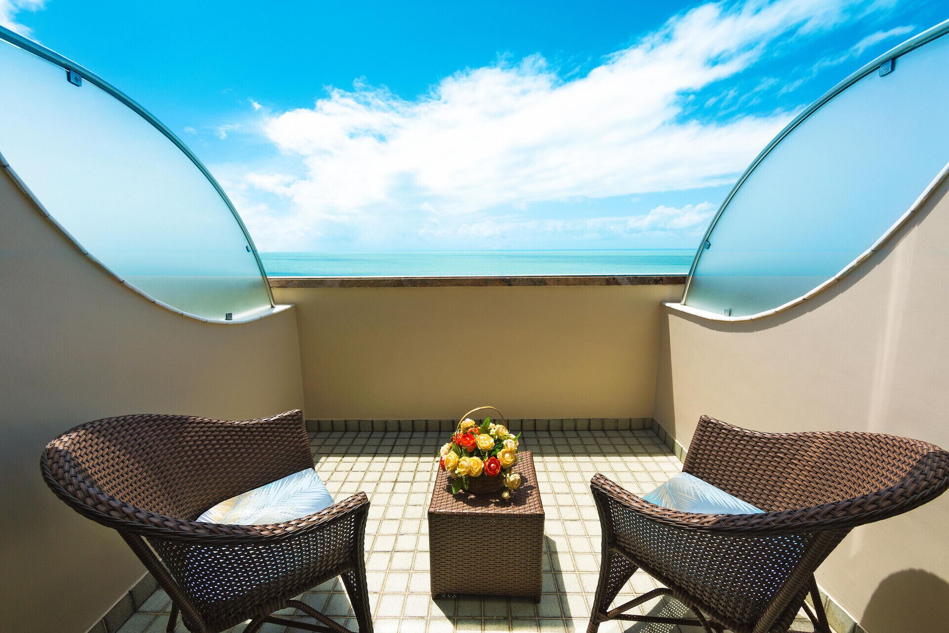 Uma varanda com duas cadeiras de vime, uma pequena mesa com um arranjo floral e vista para o oceano.