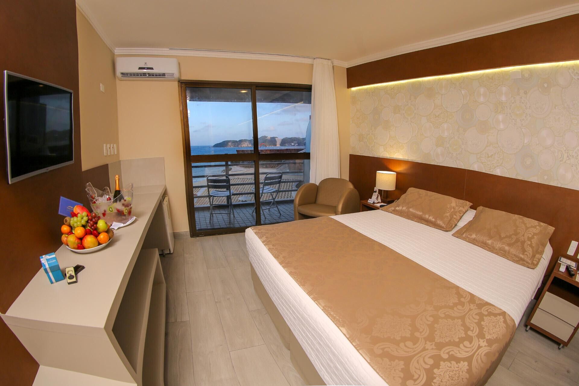 Quarto de hotel com cama, escrivaninha com fruteira, poltrona, televisão e varanda com vista.