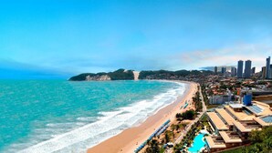 A coastal cityscape with a beach, clear blue waters, and a resort area with multiple buildings and a swimming pool. - Esmeralda Praia Hotel (Natal)