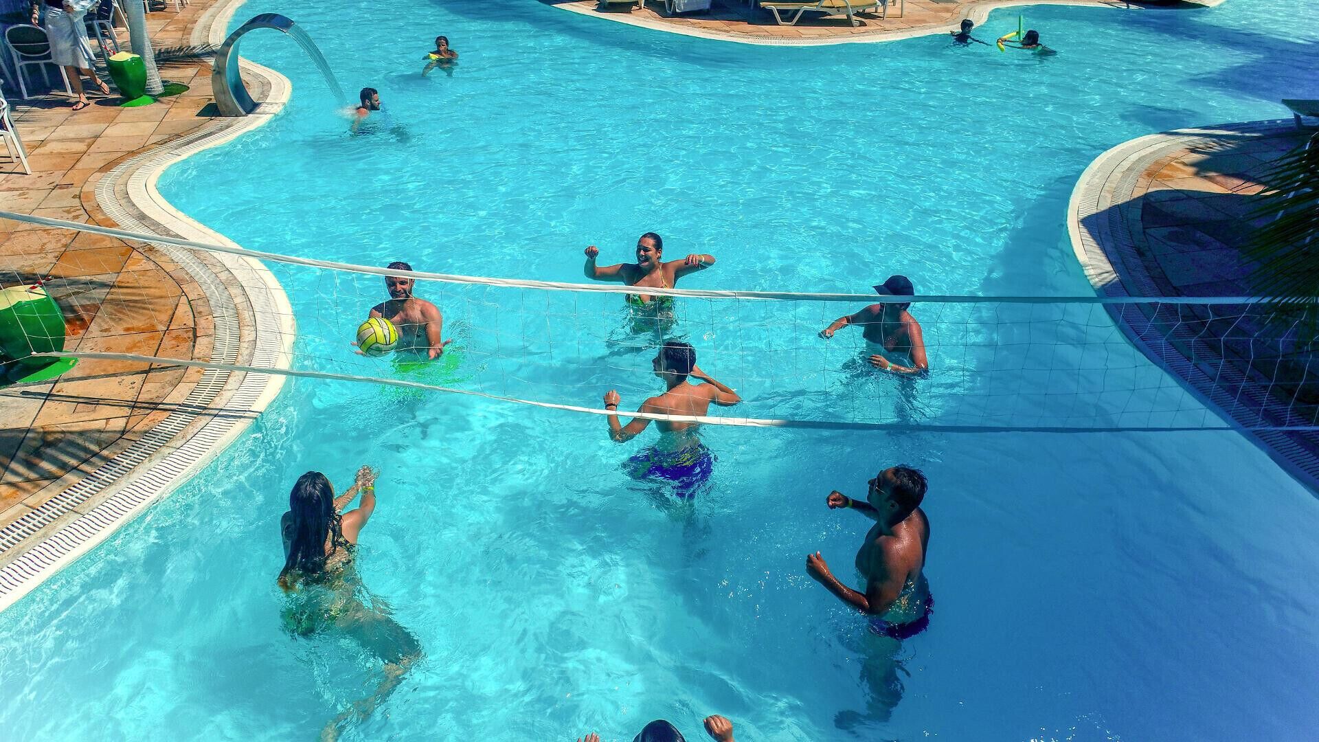 Piscina com uma rede de voleibol no centro, rodeada por espreguiçadeiras e um toldo.