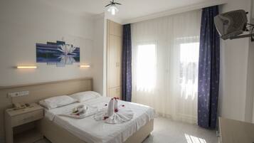 A neatly made bed with a towel and flowers, a bedside table with a phone and lamp, and a window with curtains.