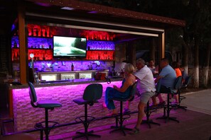 Uma área de bar com uma tela de televisão, banquetas e um balcão com garrafas e copos.