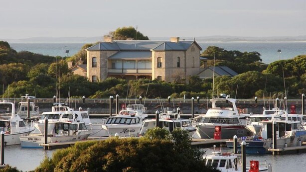 A marina with several boats docked, a stone building with a metal roof, and a view of the sea in the background.