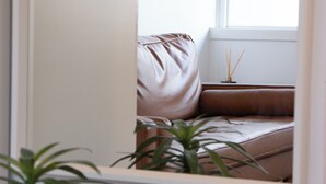 A room with a window, a brown sofa, a small table with a reed diffuser, and a potted plant.