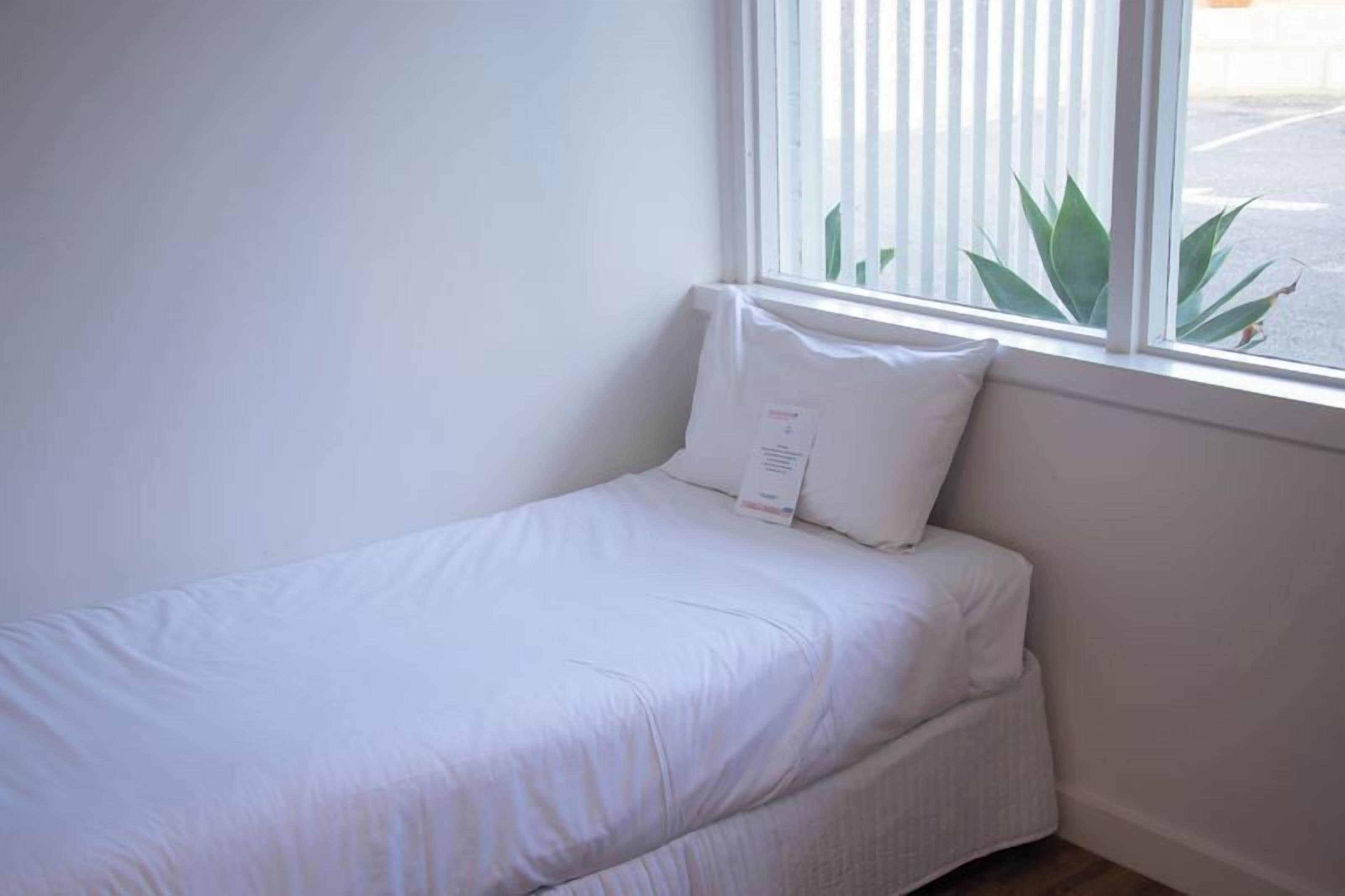 A small, neatly made bed with white bedding in a corner room with a window.