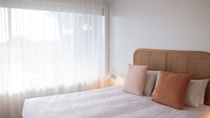A neatly made bed with white linens and two pillows, a bedside lamp, and sheer curtains on a window.