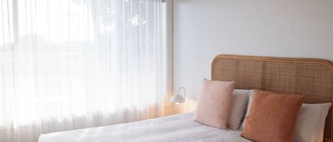 Una cama bien hecha con sábanas blancas y dos almohadas, una lámpara de noche y cortinas translúcidas en una ventana.