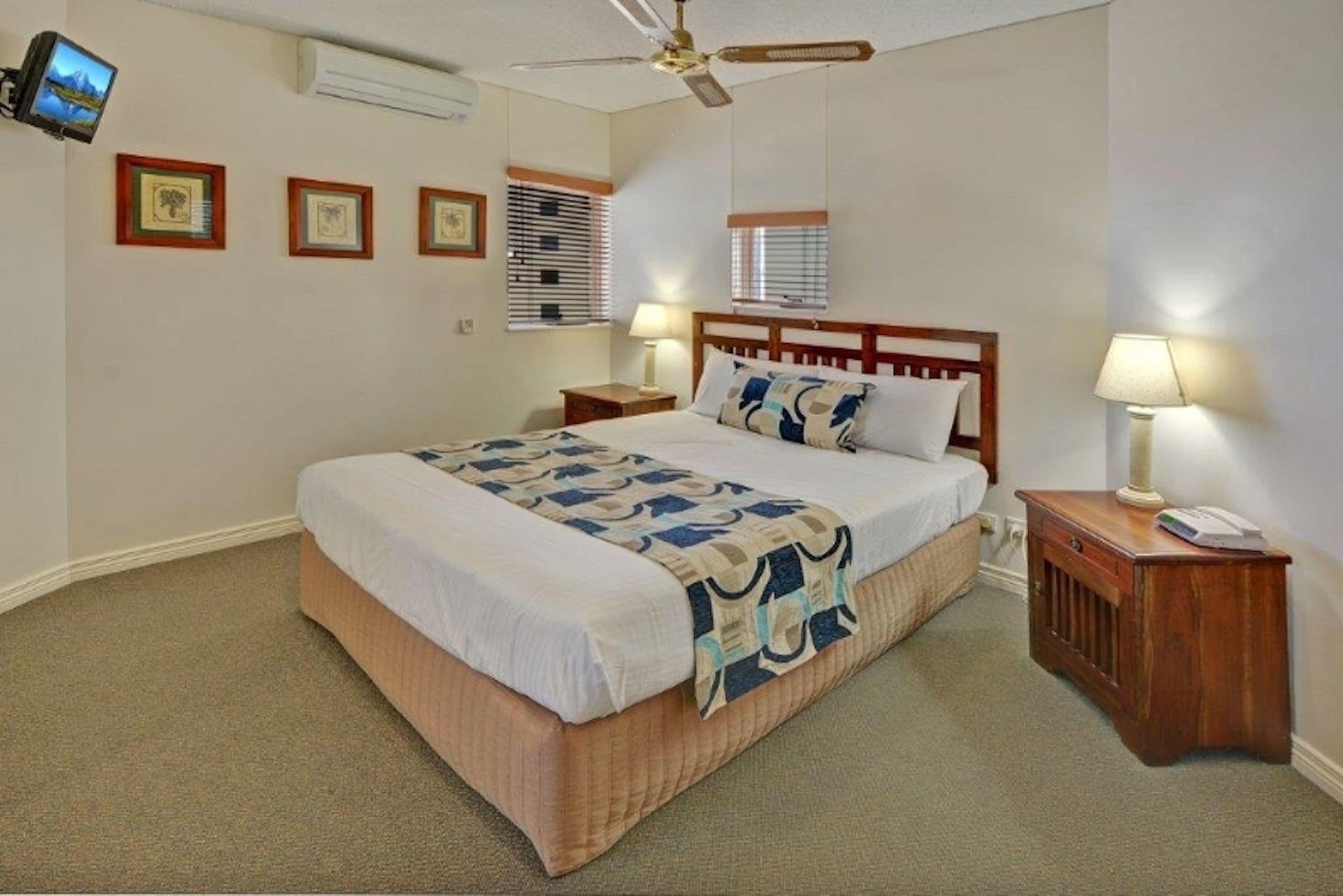 Une chambre à coucher comprenant un lit, des tables de chevet, un ventilateur de plafond, un téléviseur fixé au mur et des tableaux encadrés.