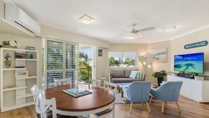 A well-lit living room with a wooden dining table, white chairs, a grey sofa, and a TV on a white cabinet. - Raffles Mooloolaba (Mooloolaba)