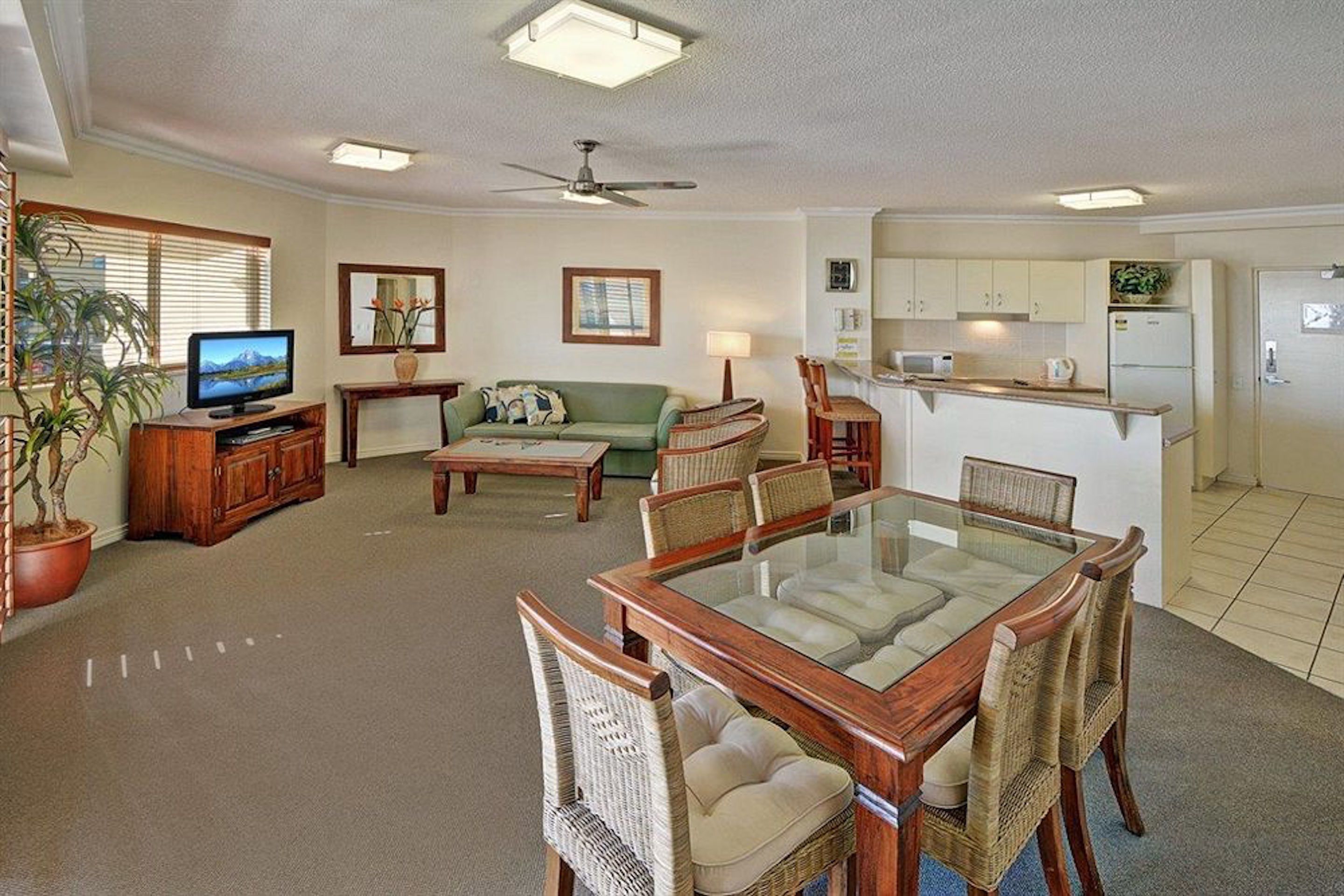 A spacious living room with a glass-top dining table, a sofa, a television, and a kitchen area.