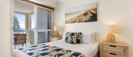 A bedroom with a bed, a nightstand, a lamp, a painting, and a view of the outdoors.