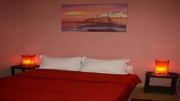 A bedroom with a bed, two bedside tables with lamps, a painting on the wall, and a red bedspread.