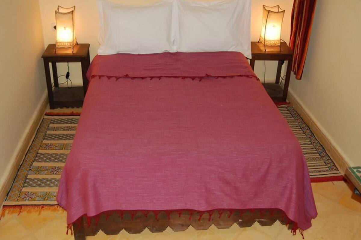 Uma cama com colcha vermelha, almofadas brancas e duas luminárias de cabeceira.