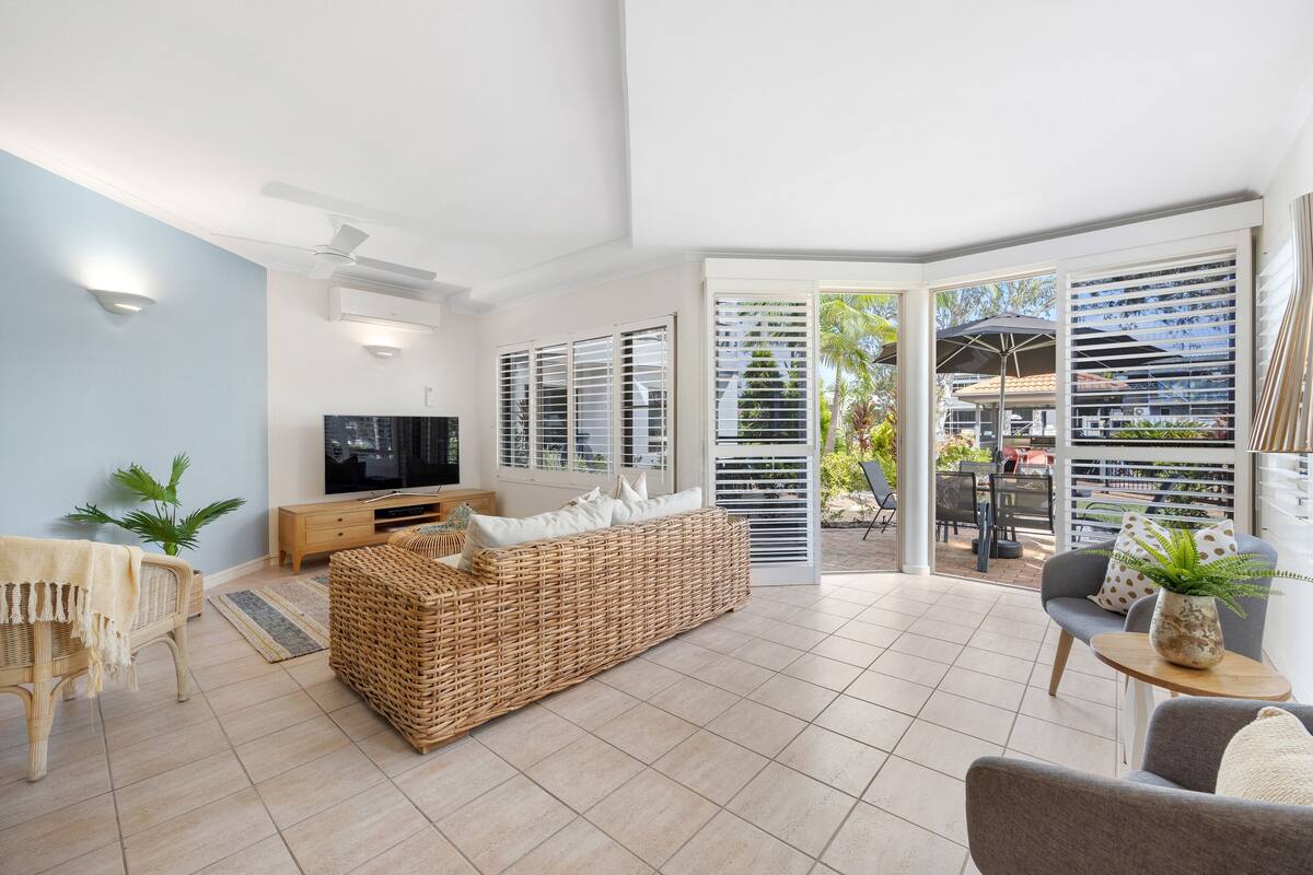 A living room with a TV, wicker sofa, and a dining area with outdoor seating.