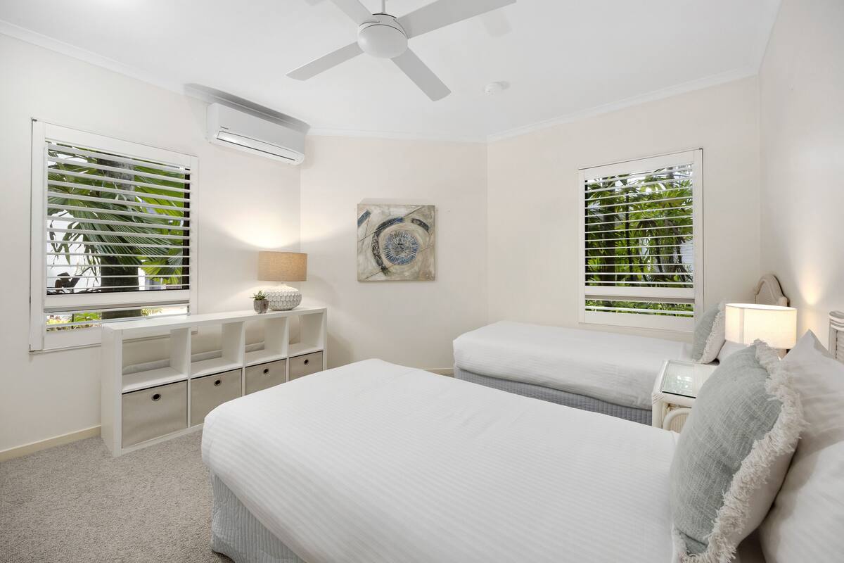 A bedroom with two beds, a ceiling fan, a nightstand with a lamp, and a window with blinds.