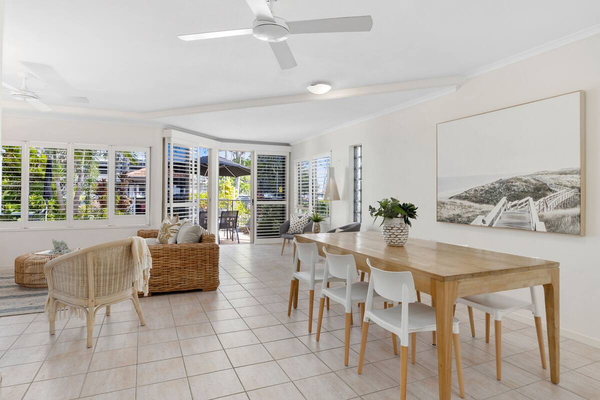 A spacious dining area with a wooden table and white chairs, a wicker sofa, and a ceiling fan.