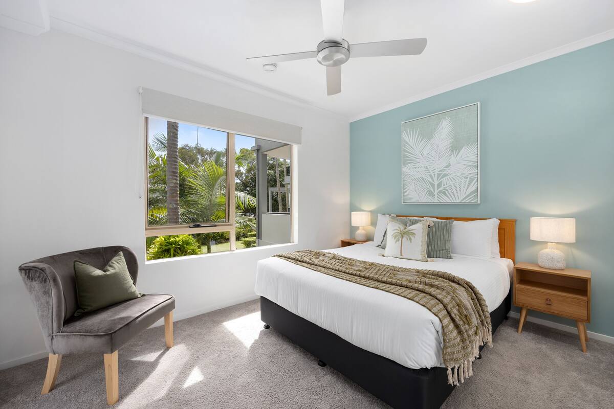 A bedroom with a bed, a chair, a nightstand, a ceiling fan, and a window with a view of greenery.