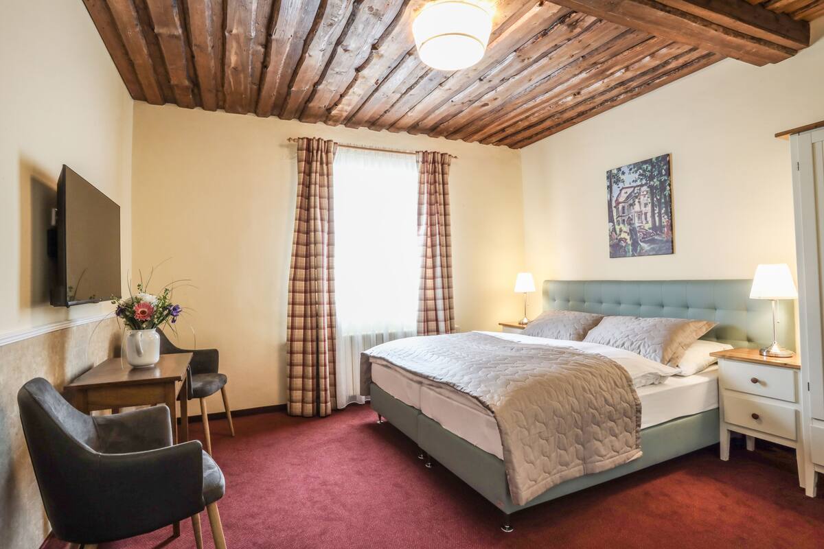 A bedroom with a bed, bedside tables, a desk with a chair, a TV mounted on the wall, and a painting on the wall.