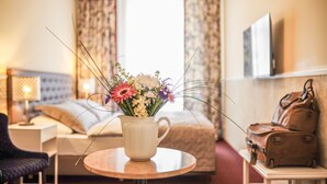 A hotel room with a bed, a small table with a vase of flowers, a chair, a nightstand with a lamp, and a suitcase on a shelf.
