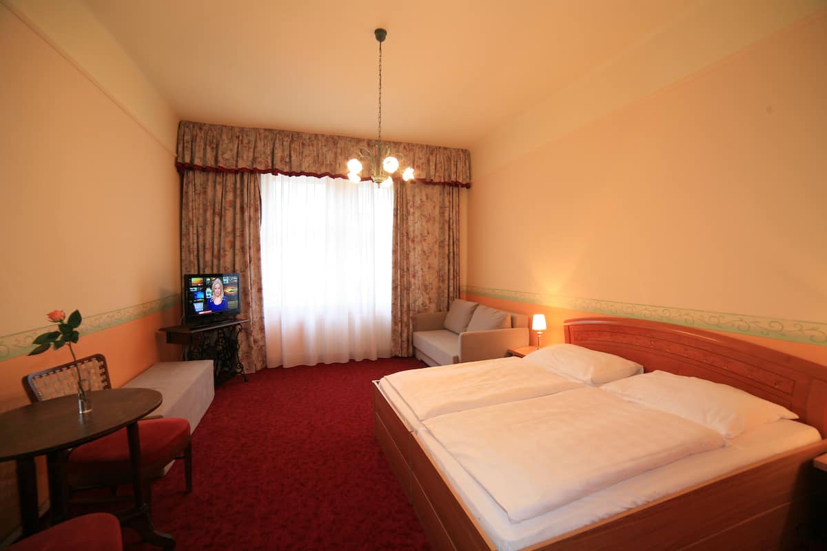 A hotel room with a large bed, a television on a small table, a desk with a chair, and a window with curtains.