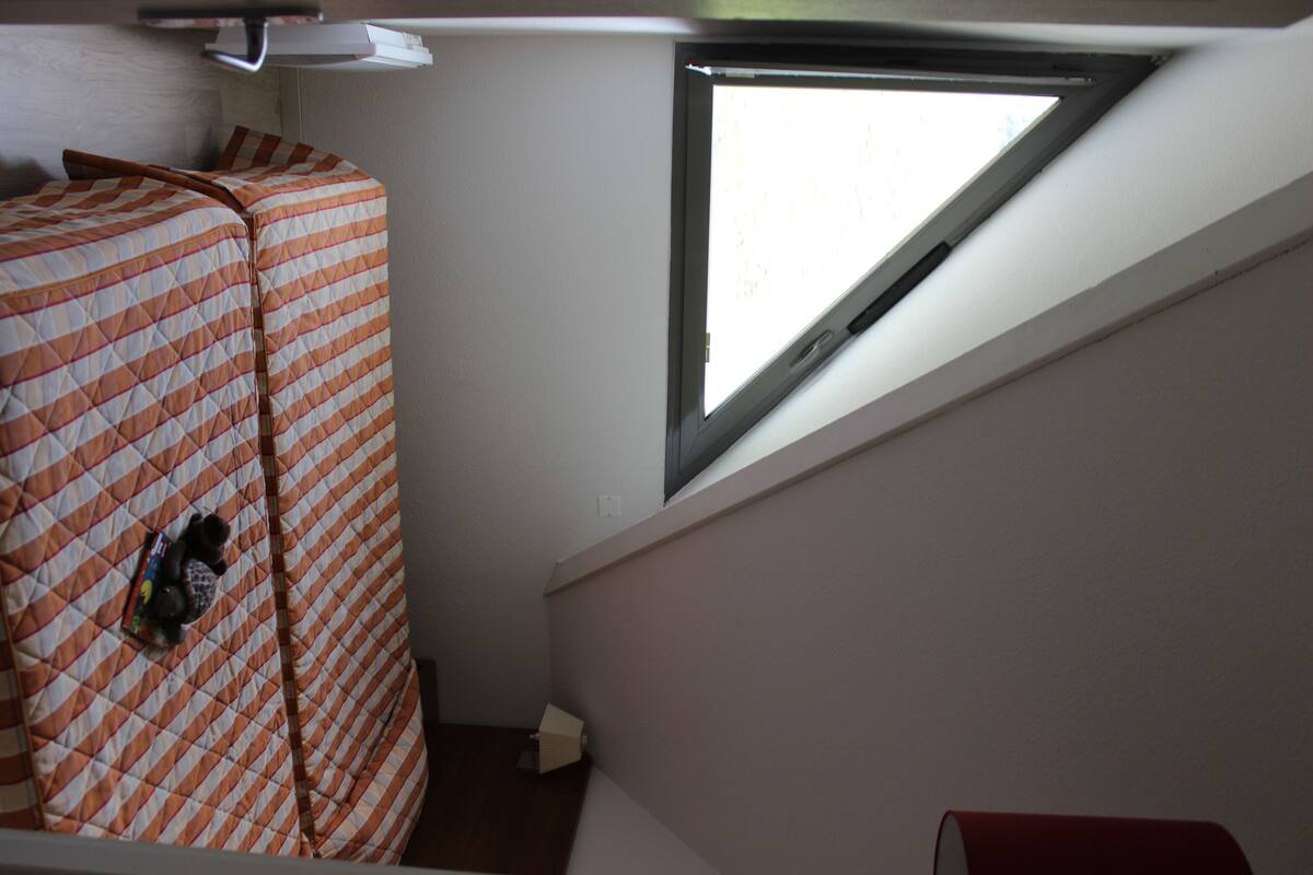 Ein Zimmer mit einem Bett, das mit einer gemusterten Decke bezogen ist, einem Fenster mit dreieckiger Aussicht und einer kleinen Schachtel auf dem Boden.