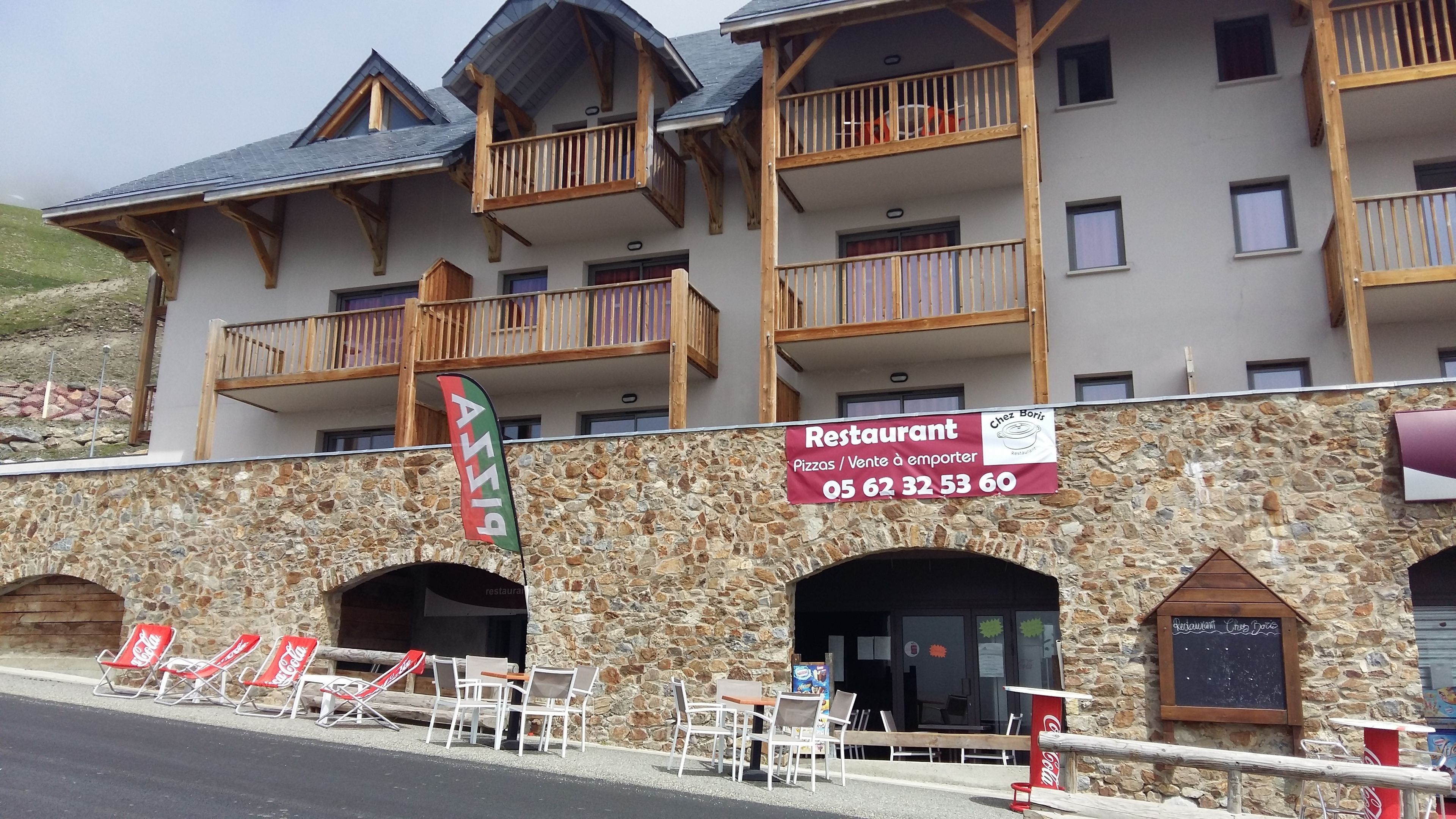 A restaurant with outdoor seating, stone facade, wooden balconies, and a sign indicating it serves pizzas and has a contact number.