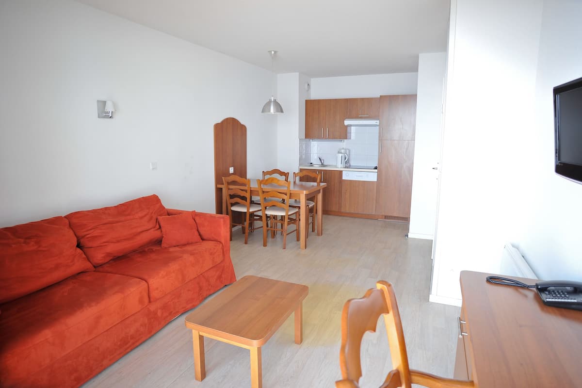 Ein modernes Wohnzimmer mit einem roten Sofa, einem Holzespeisetisch und Stühlen. Im Hintergrund ist eine Küche mit Holzschränken und einem Fernseher zu sehen.