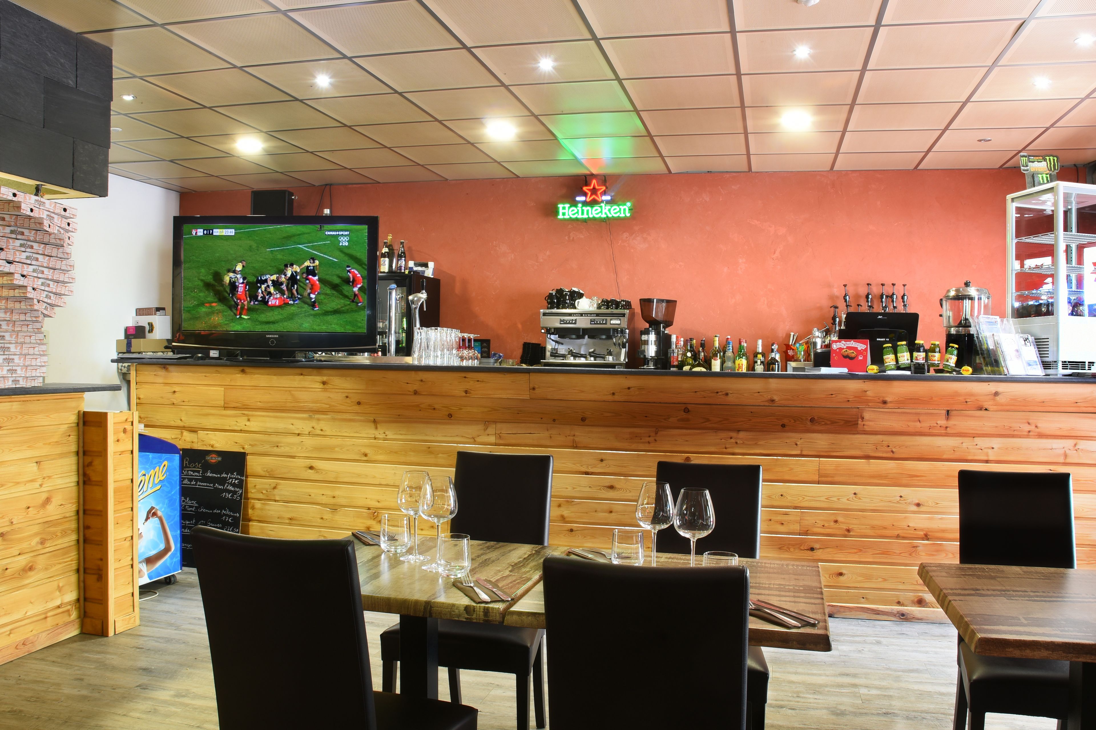 Un ristorante con arredamento in legno, un'area bar con insegna Heineken e un televisore che mostra una partita sportiva.