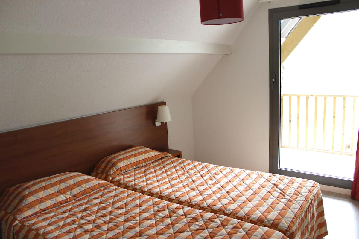 Ein Zimmer mit zwei Betten, einem Holz-Kopfstück, einem Fenster mit Blick auf einen Balkon und einer wandmontierten Lampe.
