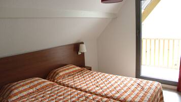 A room with two beds, a wooden headboard, a window with a view of a balcony, and a wall-mounted light fixture.