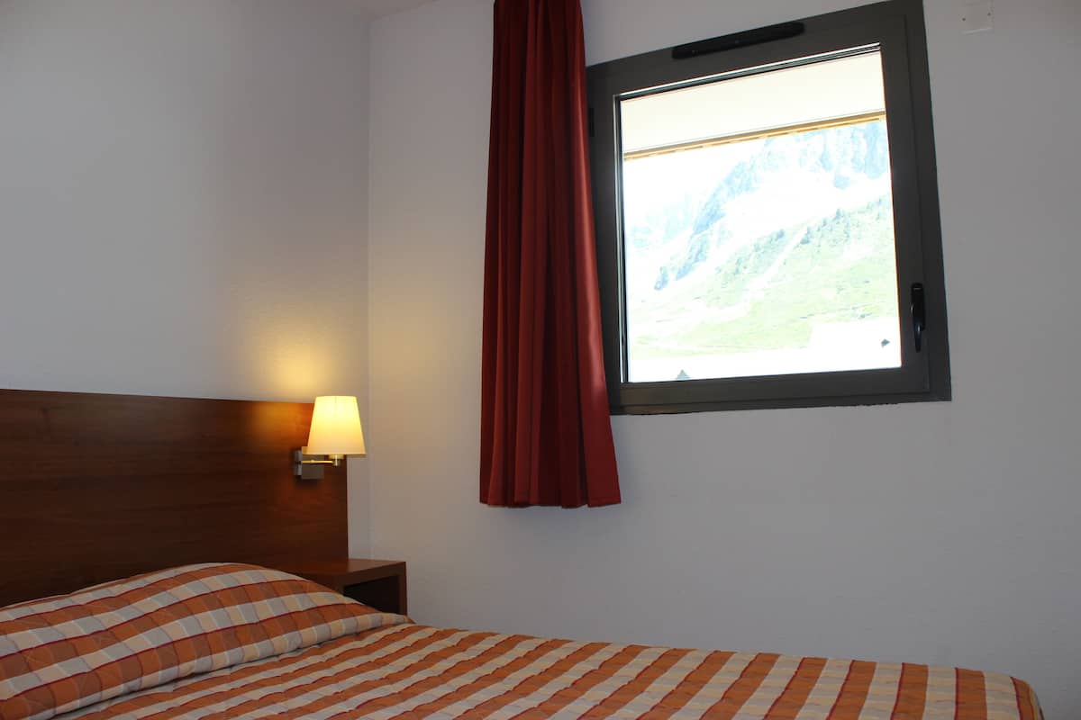Ein Hotelzimmer mit Bett, Nachttisch, Lampe, Fenster mit Bergblick und einem roten Vorhang.