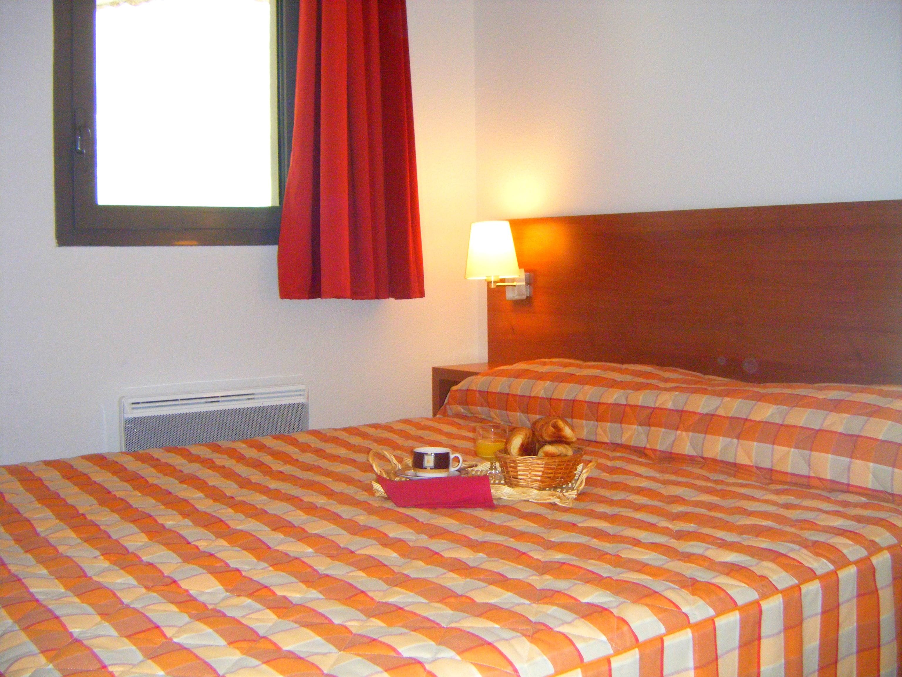 Habitación de hotel con dos camas, una ventana con cortinas rojas, una lámpara de pared y una cesta con artículos de desayuno sobre la cama.