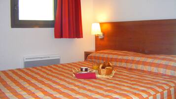 Habitación de hotel con dos camas, una ventana con cortinas rojas, una lámpara de pared y una cesta con artículos de desayuno sobre la cama.