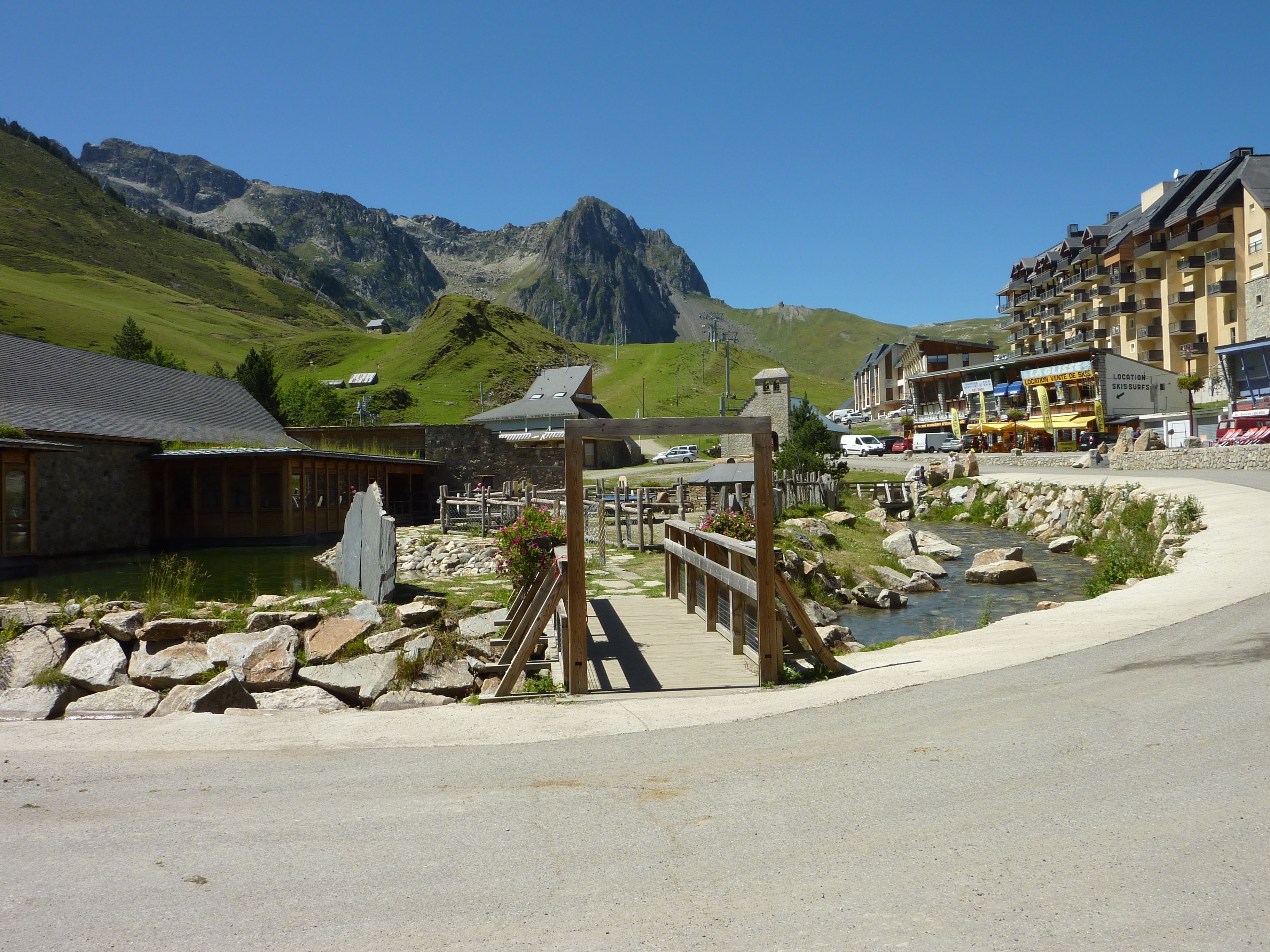 Un chalet de montagne doté d’une terrasse en bois et d’un petit étang, entouré d’un paysage rocheux et surplombant une chaîne de montagnes.
