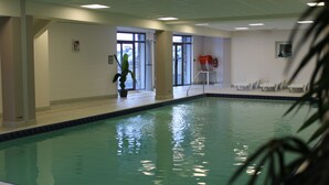 An indoor swimming pool with clear glass walls, a tiled floor, and a few chairs near the poolside. - Résidence Tourmalet (Bagnères-de-Bigorre)