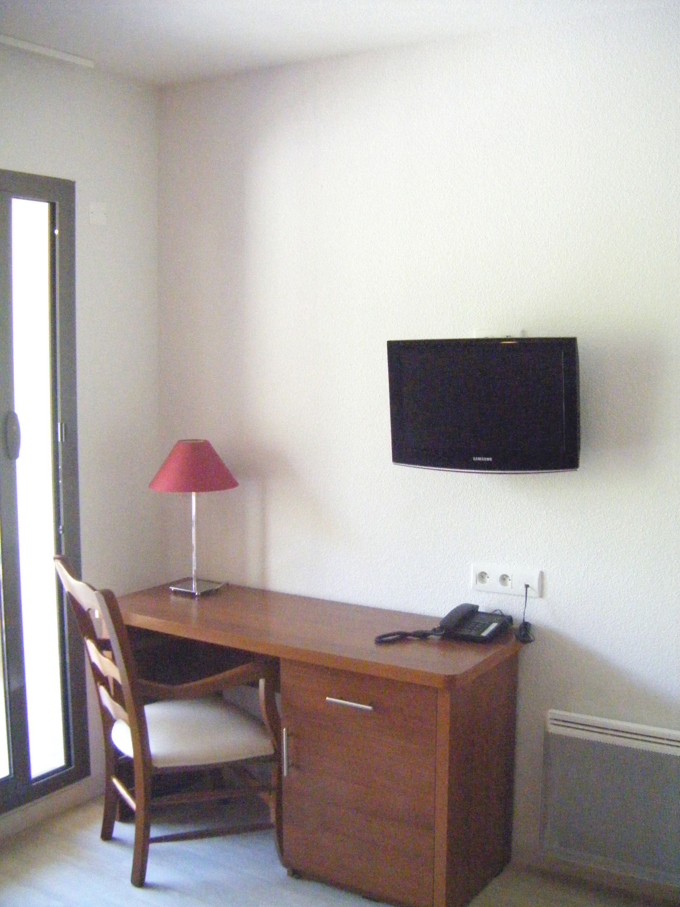 Una habitación pequeña y sencilla con un escritorio de madera, una silla, una lámpara roja, un televisor negro montado en la pared y un teléfono.