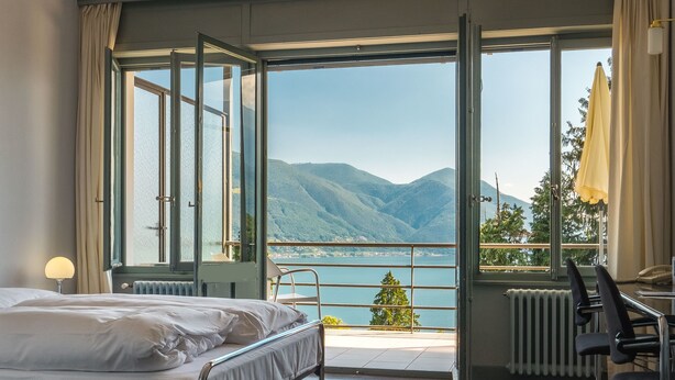 A hotel room with a bed, a desk, and a view of a lake and mountains. - Monte Verità (Ascona)