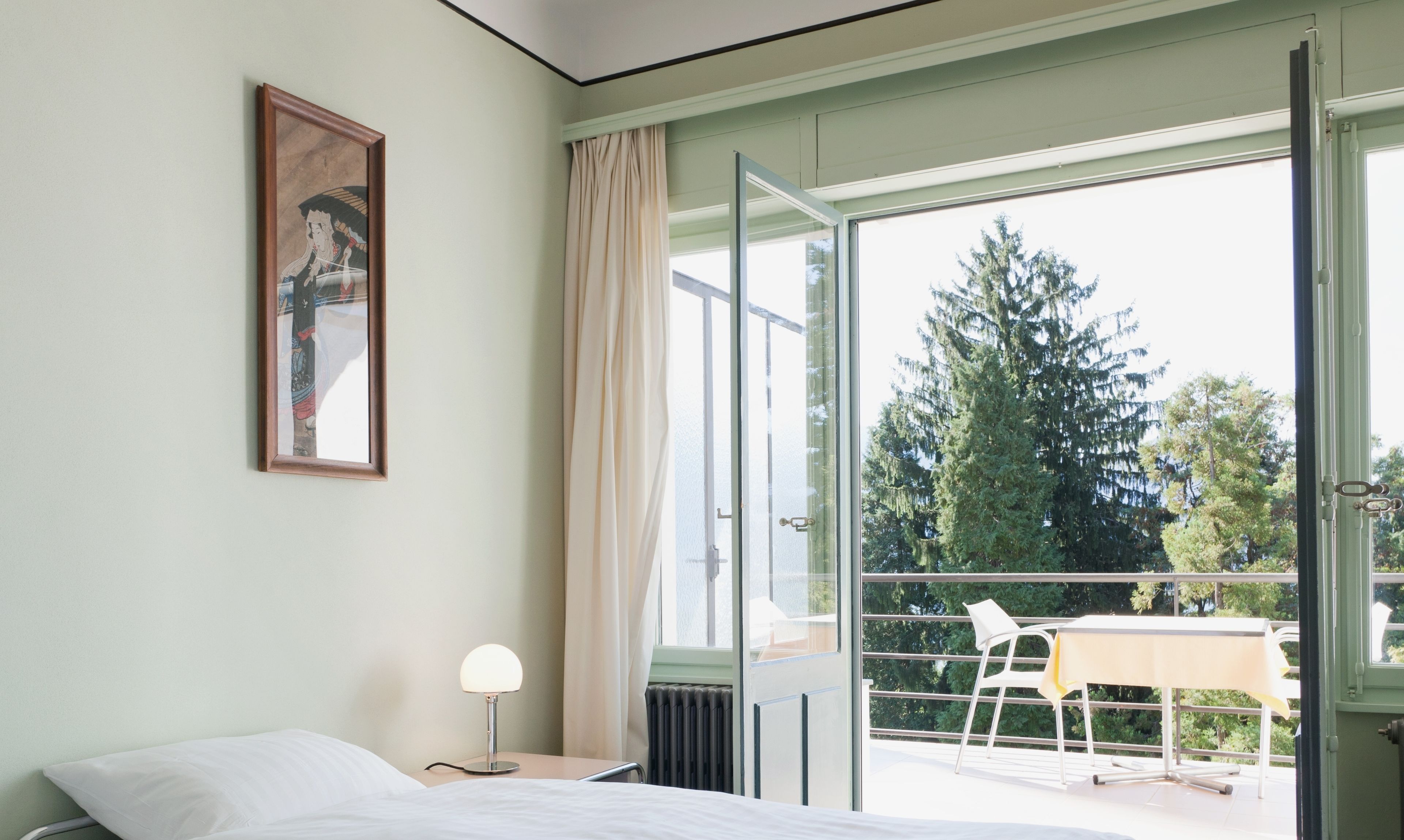 A hotel room with a bed, a bedside lamp, a painting on the wall, and a balcony with a table and chairs.