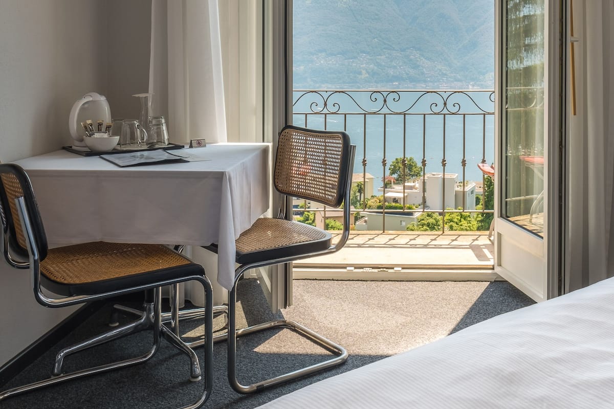 A hotel room with a bed, a table with a chair, a view of mountains through a balcony.