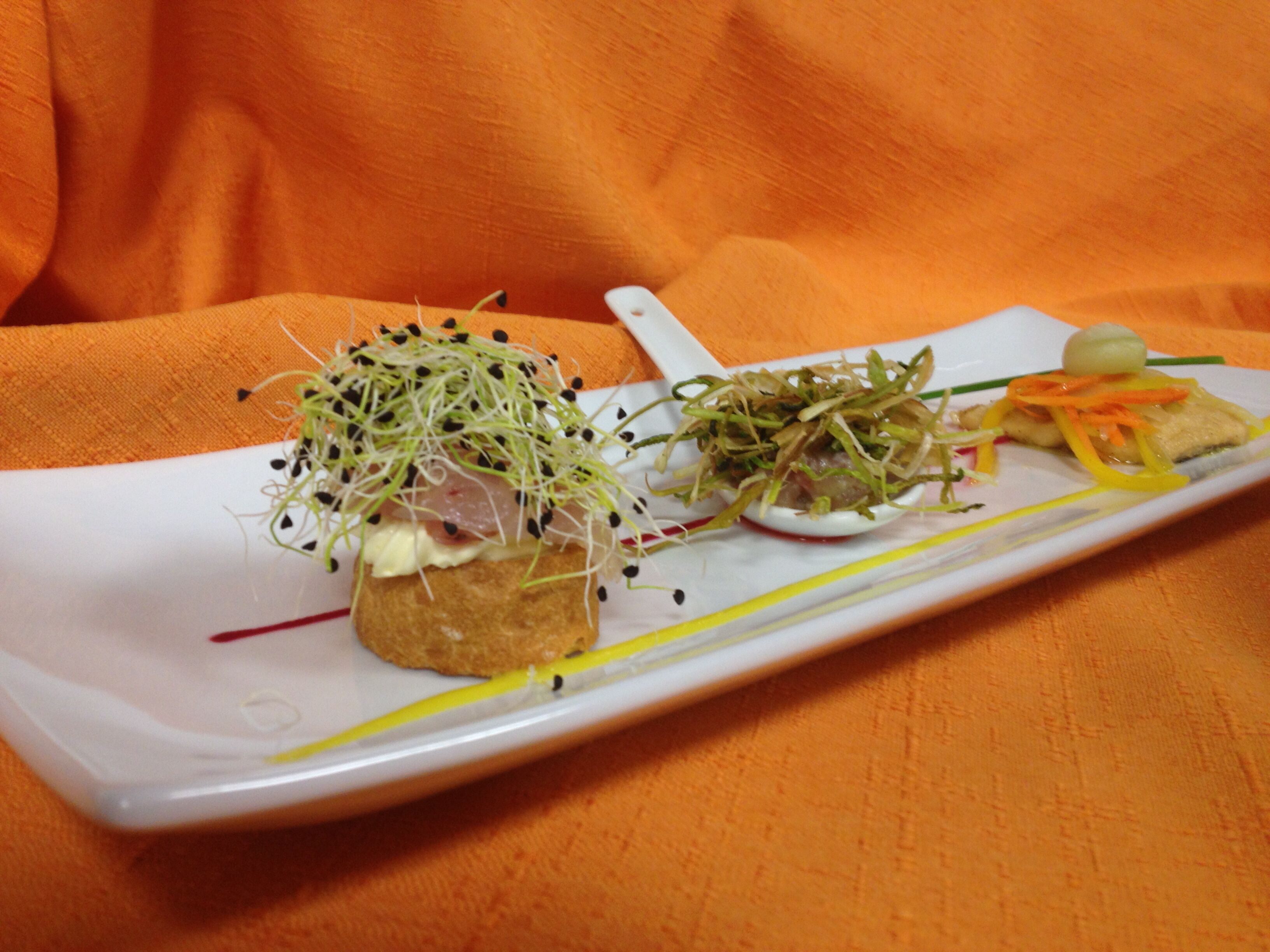 Plato blanco rectangular con varios alimentos, incluyendo un trozo de pan cubierto de brotes y una pequeña cucharada de crema, sobre una superficie naranja texturizada.