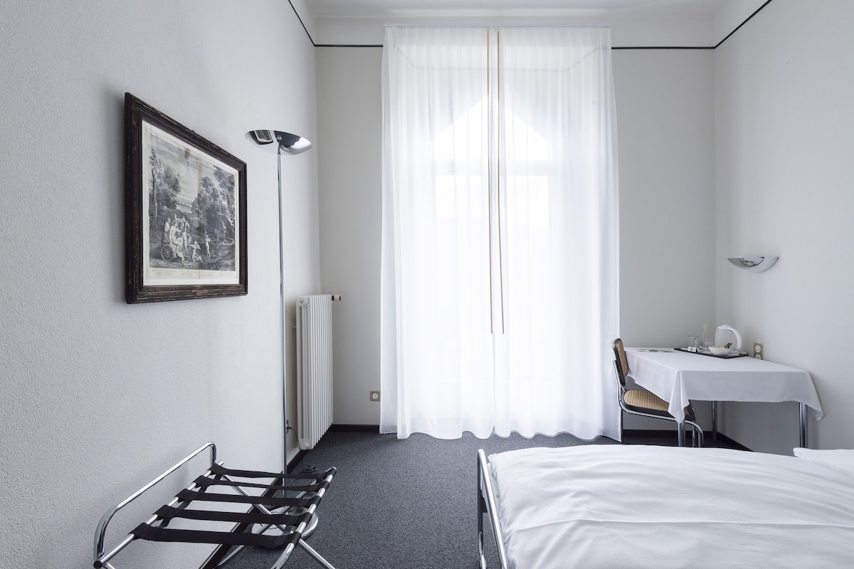 A hotel room with a bed, a chair, a table, a radiator, a window with curtains, and a framed picture on the wall.