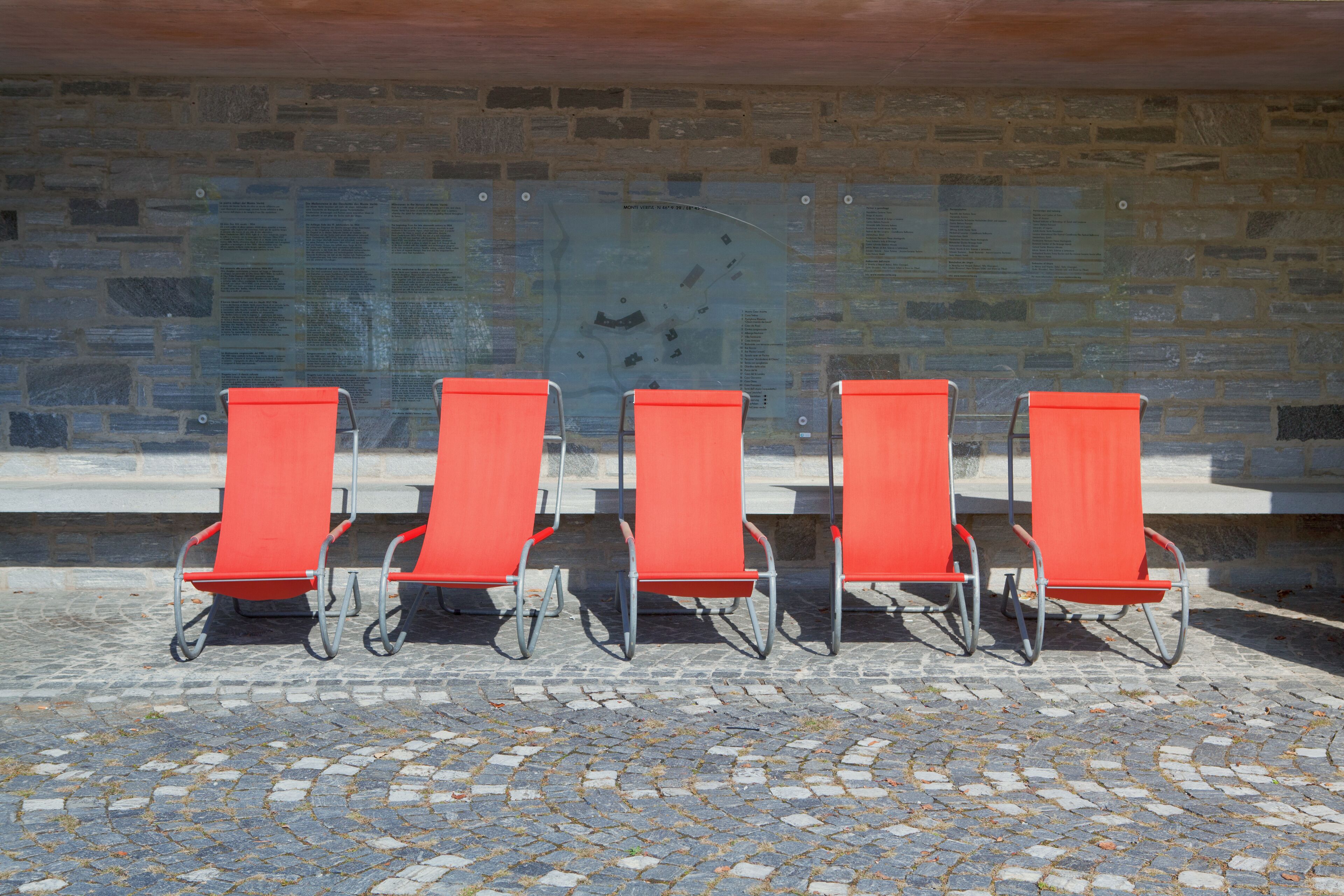 A row of five orange chairs with metal frames, arranged on a cobblestone pavement against a stone wall with informational plaques.