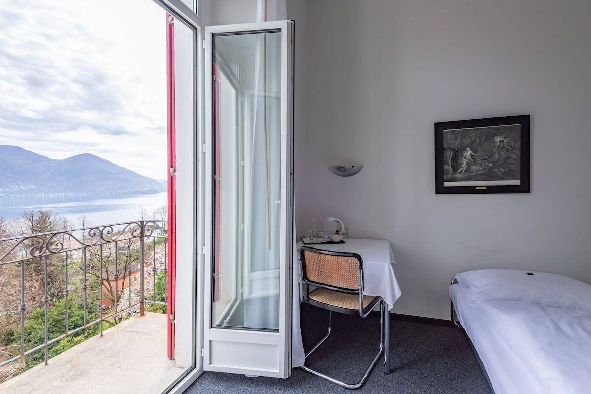 A room with a balcony, a bed, a chair, a table, and a framed picture on the wall.