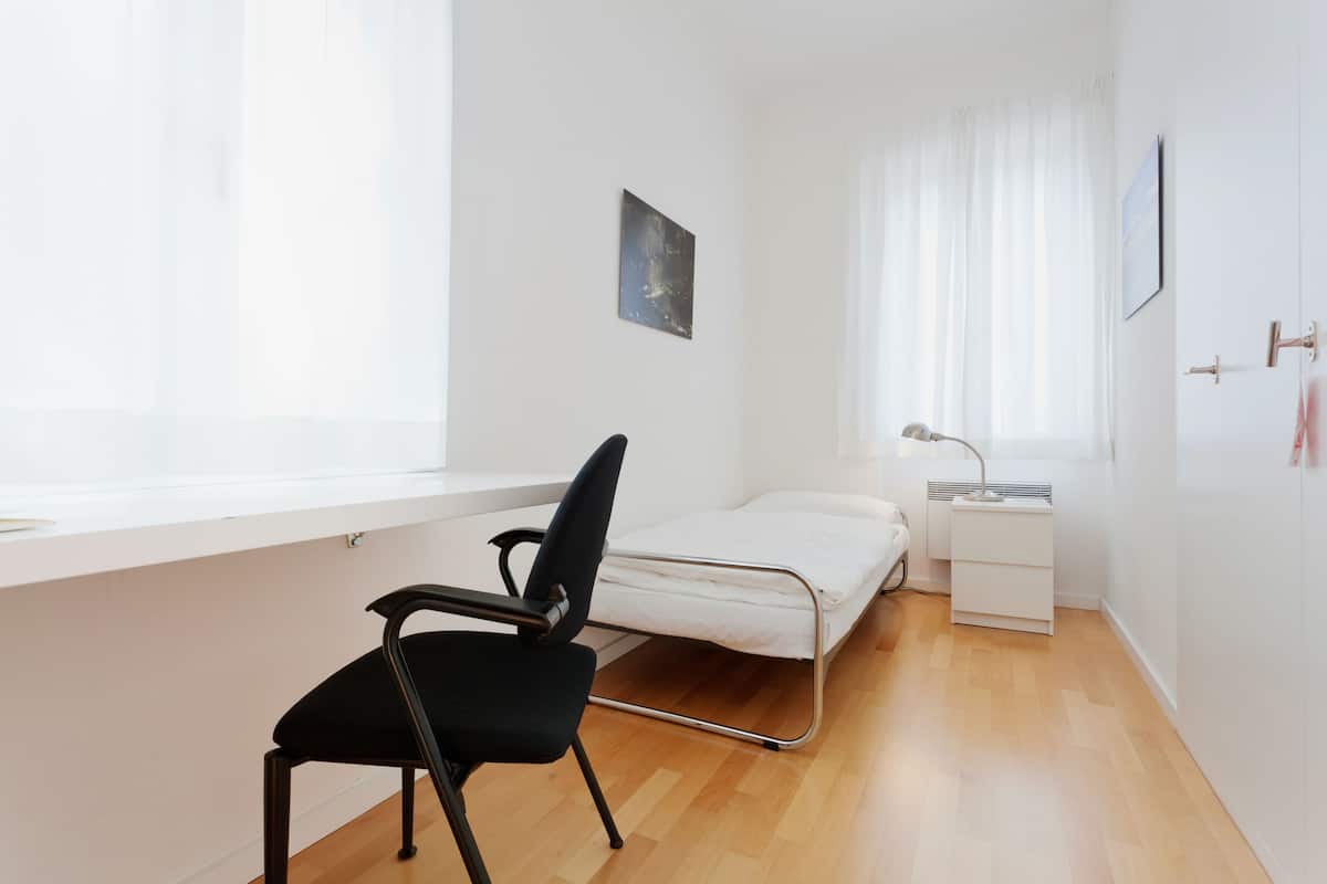 A small, bright room with a single bed, a desk, a chair, and a window with sheer curtains.