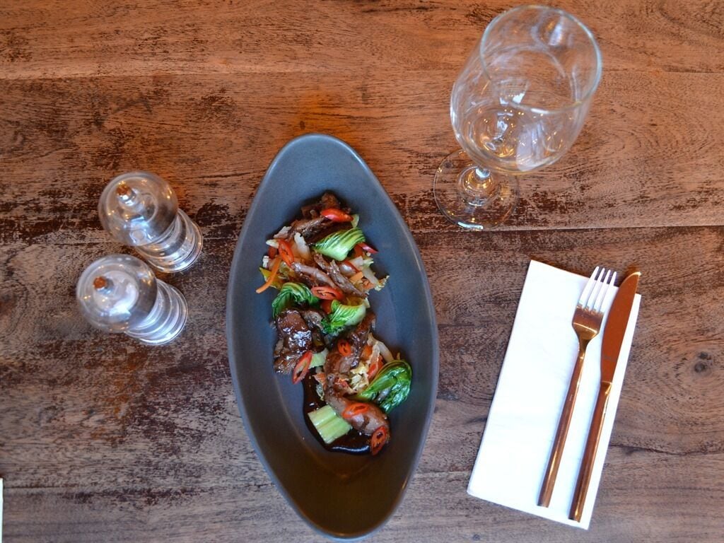 A meal on a dark plate with a fork and knife on a napkin, salt and pepper shakers, and a glass of water on a wooden table.