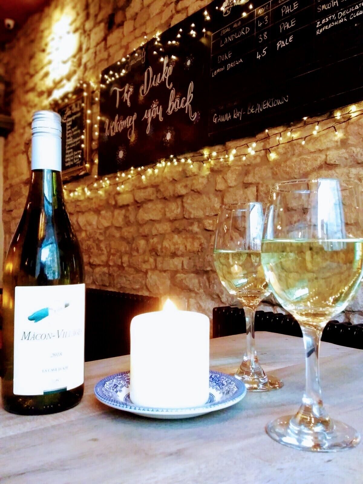A bottle of Macon-Vill wine, a lit candle, and two glasses of white wine on a table with a stone wall and a menu in the background.